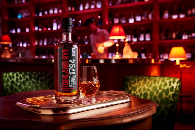 a bottle of highland rye whisky with a dram on the rocks in a lounge bar setting with tables chairs and a bar in the background