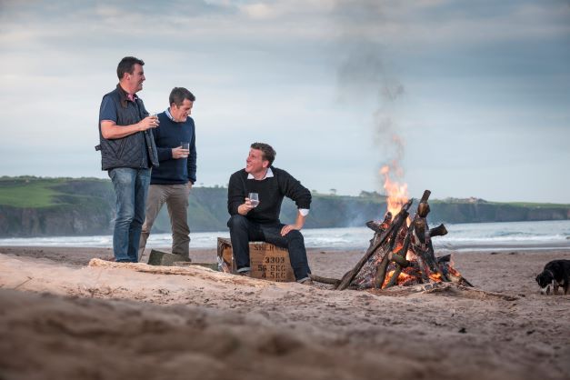 three men drinking on the beach keeping warm by a wood fire