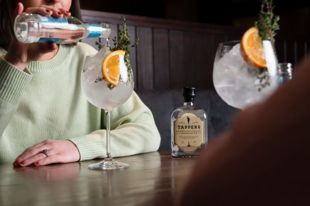 a gin and tonic being poured at a wooden table with citrus garnish and a bottle of gin on the table